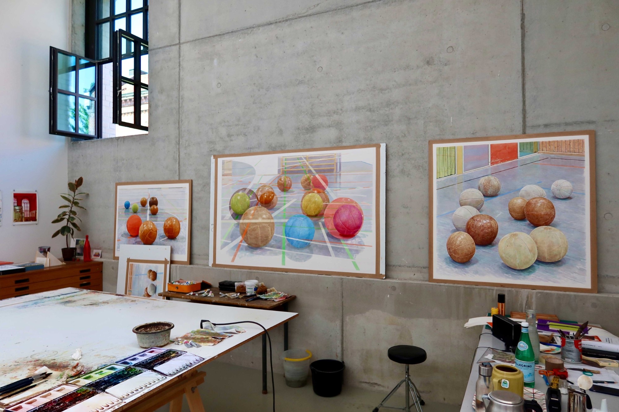 Blick in das Atelier: Drei große Aquarelle mit Bällen in Hallen lehnen an einer Betonwand, davor ein Arbeitstisch mit Farbkästen und Pinseln, links ein hohes Fenster und ein Gummibaum.
